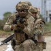 Two heavily equipped military or special operations personnel are shown in a tactical formation, with the foreground soldier aiming a rifle directly toward the camera. Both individuals wear multicam camouflage uniforms, tactical plate carriers, and ballistic helmets with accessory mounts. The setting appears to be an outdoor training or operational environment with an industrial structure visible in the background.
