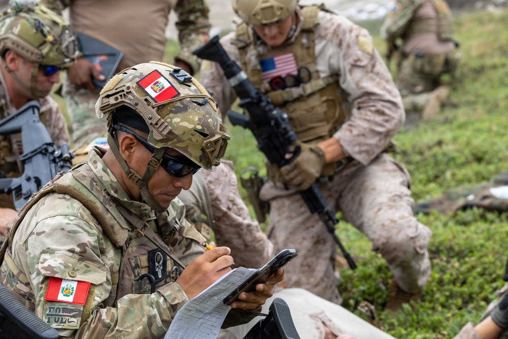 The image shows a joint military exercise or operation involving Peruvian and American armed forces personnel. A Peruvian soldier in the foreground is writing on a clipboard while holding a tablet device, suggesting coordination or documentation activities. Behind him, a U.S. Marine in MARPAT desert camouflage carries an assault rifle and observes the surroundings.