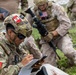 The image shows a joint military exercise or operation involving Peruvian and American armed forces personnel. A Peruvian soldier in the foreground is writing on a clipboard while holding a tablet device, suggesting coordination or documentation activities. Behind him, a U.S. Marine in MARPAT desert camouflage carries an assault rifle and observes the surroundings.
