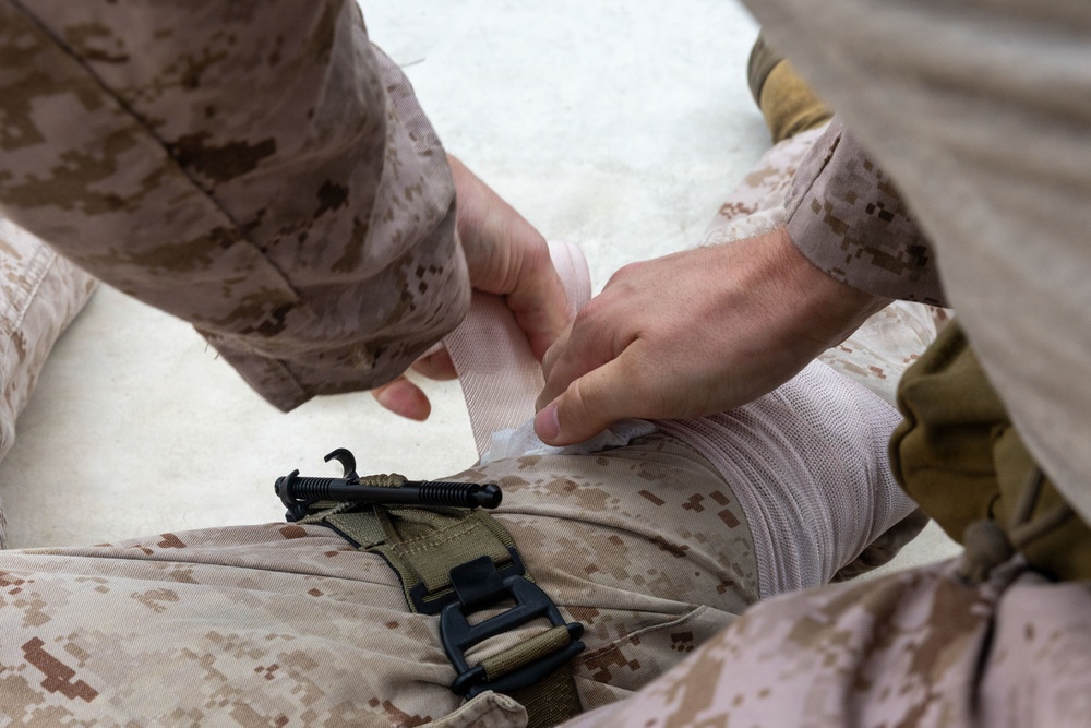 The image shows a person in military desert camouflage (MARPAT pattern) applying a white bandage or compression wrap to what appears to be a leg or limb of another individual also wearing the same camouflage uniform. A tourniquet or restraining strap with a black buckle and hook mechanism is visible on the casualty's leg. This appears to depict a tactical combat casualty care (TCCC) or first aid scenario.