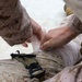 The image shows a person in military desert camouflage (MARPAT pattern) applying a white bandage or compression wrap to what appears to be a leg or limb of another individual also wearing the same camouflage uniform. A tourniquet or restraining strap with a black buckle and hook mechanism is visible on the casualty's leg. This appears to depict a tactical combat casualty care (TCCC) or first aid scenario.