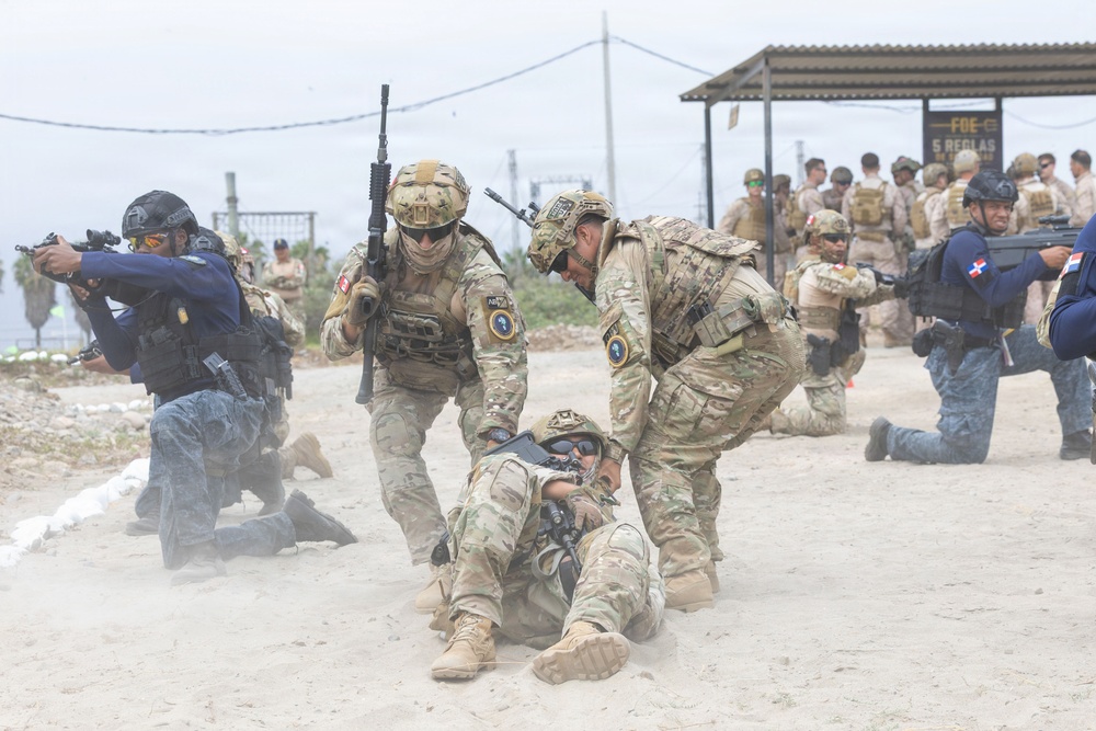 The image depicts a military or law enforcement training exercise taking place on a sandy outdoor terrain. Multiple personnel in various uniforms and tactical gear are engaged in a simulated casualty evacuation and tactical response drill. A sign in the background reading 'FOE 5 REGLAS' suggests a Spanish-speaking military or security organization context.