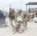 The image depicts a military or law enforcement training exercise taking place on a sandy outdoor terrain. Multiple personnel in various uniforms and tactical gear are engaged in a simulated casualty evacuation and tactical response drill. A sign in the background reading 'FOE 5 REGLAS' suggests a Spanish-speaking military or security organization context.