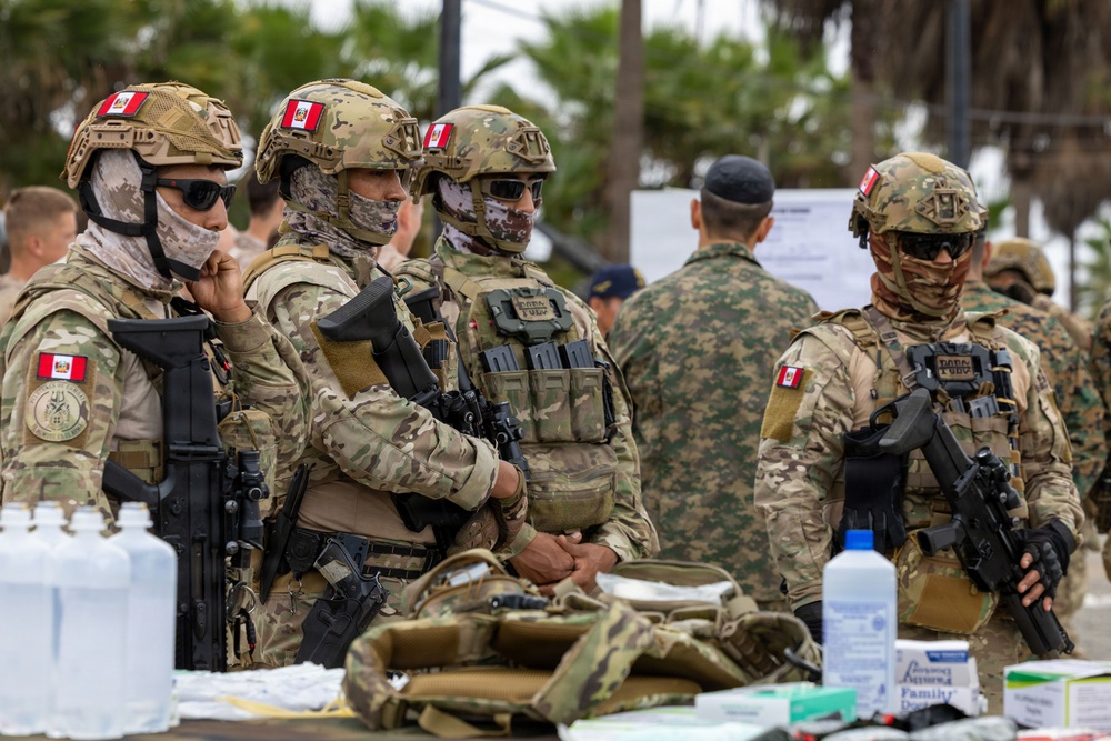 The image shows a group of heavily armed military personnel wearing camouflage uniforms and tactical gear, with Peruvian flag patches visible on their helmets and shoulders. They appear to be gathered at an outdoor staging area with medical supplies and equipment on a table in the foreground. The scene suggests a military operation, humanitarian mission, or joint exercise.