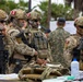 The image shows a group of heavily armed military personnel wearing camouflage uniforms and tactical gear, with Peruvian flag patches visible on their helmets and shoulders. They appear to be gathered at an outdoor staging area with medical supplies and equipment on a table in the foreground. The scene suggests a military operation, humanitarian mission, or joint exercise.