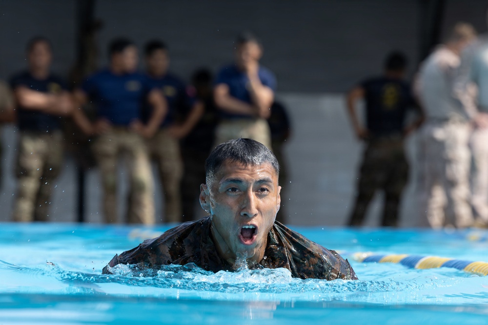 A military service member in camouflage uniform is swimming in a pool, mouth open as if exerting effort or calling out. Observers in military uniforms and civilian attire stand watching in the blurred background. This appears to be a military water survival or combat swimmer training exercise.