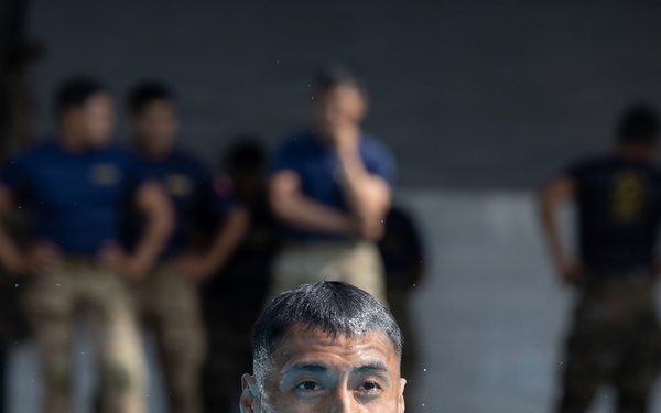 A military service member in camouflage uniform is swimming in a pool, mouth open as if exerting effort or calling out. Observers in military uniforms and civilian attire stand watching in the blurred background. This appears to be a military water survival or combat swimmer training exercise.