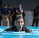 A military service member in camouflage uniform is swimming in a pool, mouth open as if exerting effort or calling out. Observers in military uniforms and civilian attire stand watching in the blurred background. This appears to be a military water survival or combat swimmer training exercise.