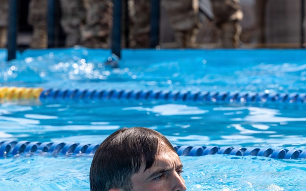 A person is swimming in an outdoor pool with blue lane dividers visible. The swimmer's head is above water, appearing to be performing a low-profile swim stroke. In the background, individuals wearing military-style camouflage uniforms are visible, suggesting this may be a military training or fitness assessment.