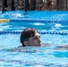 A person is swimming in an outdoor pool with blue lane dividers visible. The swimmer's head is above water, appearing to be performing a low-profile swim stroke. In the background, individuals wearing military-style camouflage uniforms are visible, suggesting this may be a military training or fitness assessment.