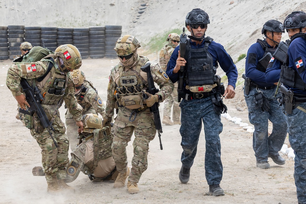The image depicts a military or law enforcement training exercise involving personnel in two distinct uniforms: multicam/camouflage battle dress and dark blue tactical gear. Several soldiers in camouflage are performing a casualty evacuation drill, dragging a simulated casualty across a dirt training ground. Personnel in dark blue uniforms with what appear to be Dominican Republic flag patches are observing or participating alongside the camouflage-clad soldiers.