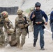 The image depicts a military or law enforcement training exercise involving personnel in two distinct uniforms: multicam/camouflage battle dress and dark blue tactical gear. Several soldiers in camouflage are performing a casualty evacuation drill, dragging a simulated casualty across a dirt training ground. Personnel in dark blue uniforms with what appear to be Dominican Republic flag patches are observing or participating alongside the camouflage-clad soldiers.