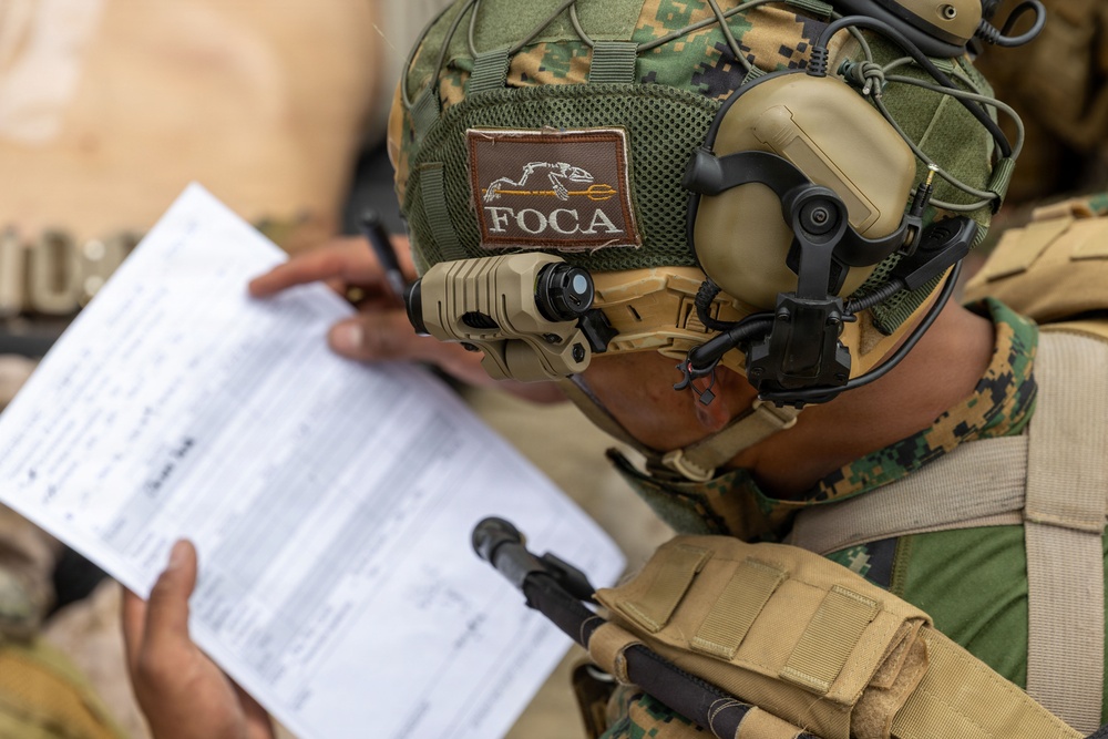 A military personnel wearing MARPAT (Marine digital camouflage) gear and a tactical helmet is reviewing a printed document. The helmet bears a 'FOCA' patch with a skeletal animal logo, and the soldier is equipped with advanced communications gear including a headset and helmet-mounted device. The image is taken from a close overhead angle, focusing on the helmet and the document being reviewed.