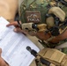 A military personnel wearing MARPAT (Marine digital camouflage) gear and a tactical helmet is reviewing a printed document. The helmet bears a 'FOCA' patch with a skeletal animal logo, and the soldier is equipped with advanced communications gear including a headset and helmet-mounted device. The image is taken from a close overhead angle, focusing on the helmet and the document being reviewed.