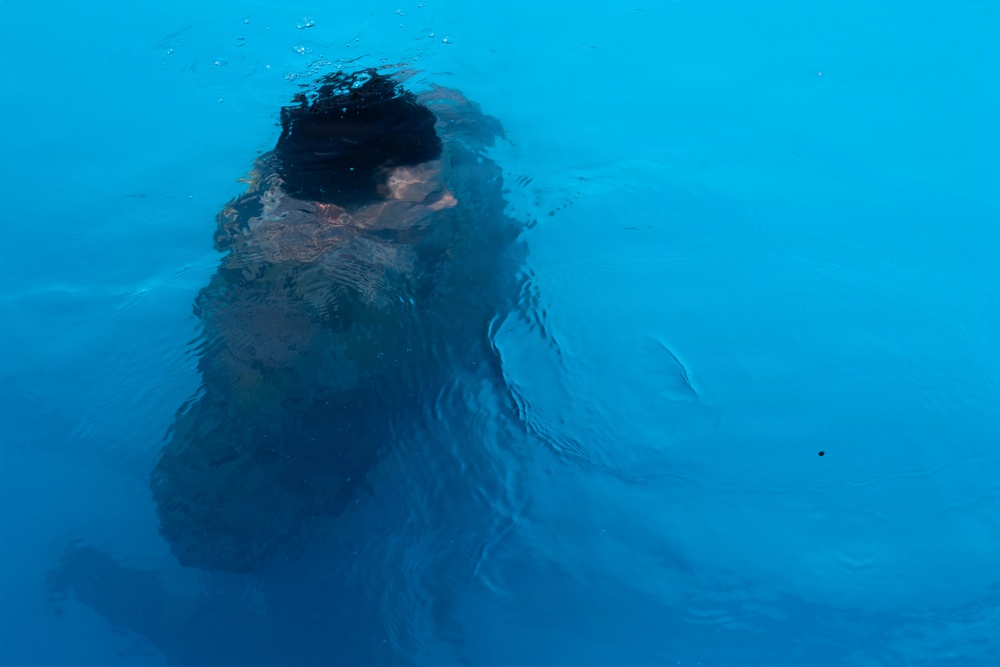 The image shows a person submerged or partially submerged in bright blue water, viewed from above. The figure appears to be swimming or diving, with dark hair visible at the surface. The water's refractive distortion creates an abstract, impressionistic quality to the image.