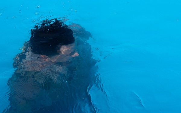 The image shows a person submerged or partially submerged in bright blue water, viewed from above. The figure appears to be swimming or diving, with dark hair visible at the surface. The water's refractive distortion creates an abstract, impressionistic quality to the image.