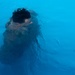 The image shows a person submerged or partially submerged in bright blue water, viewed from above. The figure appears to be swimming or diving, with dark hair visible at the surface. The water's refractive distortion creates an abstract, impressionistic quality to the image.