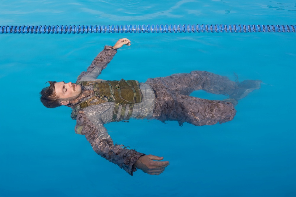 A man in full tactical/military-style clothing including a vest or plate carrier is floating face-up on the surface of a blue swimming pool. His eyes are closed and his arms are spread outward. The image appears to be a staged or artistic/training photograph taken from directly above.
