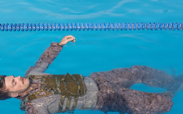 A man in full tactical/military-style clothing including a vest or plate carrier is floating face-up on the surface of a blue swimming pool. His eyes are closed and his arms are spread outward. The image appears to be a staged or artistic/training photograph taken from directly above.