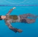 A man in full tactical/military-style clothing including a vest or plate carrier is floating face-up on the surface of a blue swimming pool. His eyes are closed and his arms are spread outward. The image appears to be a staged or artistic/training photograph taken from directly above.