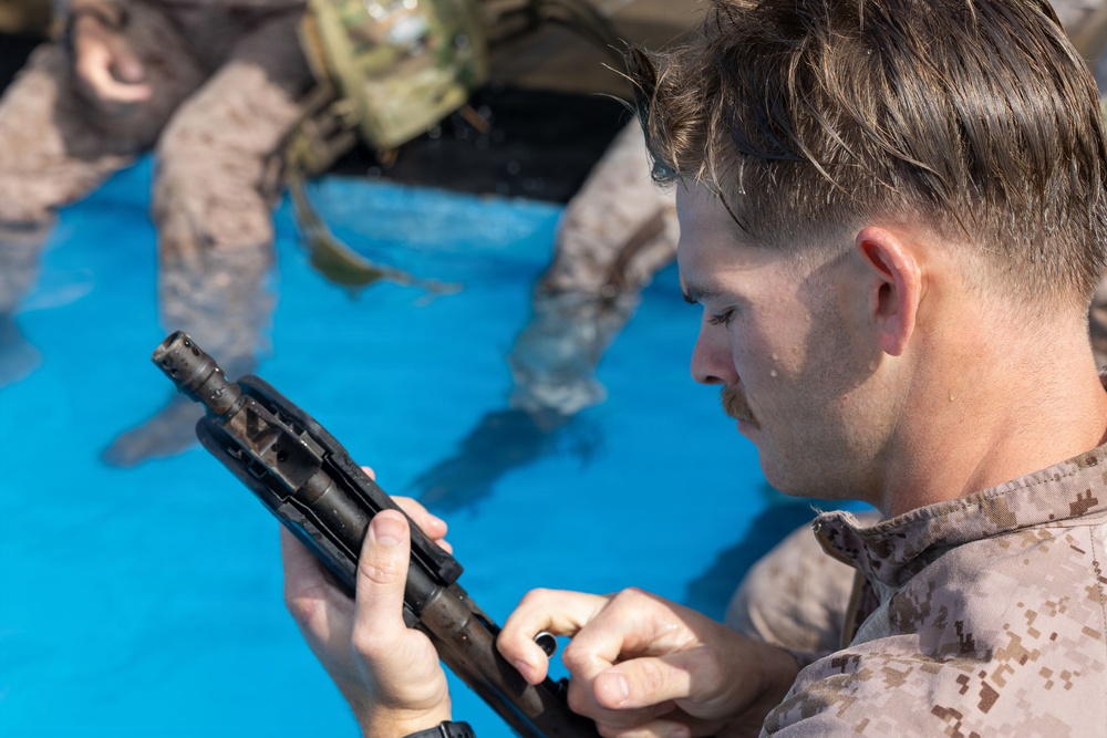 A military service member in desert MARPAT camouflage uniform is inspecting or field-stripping a firearm component while partially submerged in water, likely during a water survival or amphibious training exercise. A second service member in similar uniform is visible in the blurred background, also in the water. The setting appears to be a training pool based on the bright blue water visible.