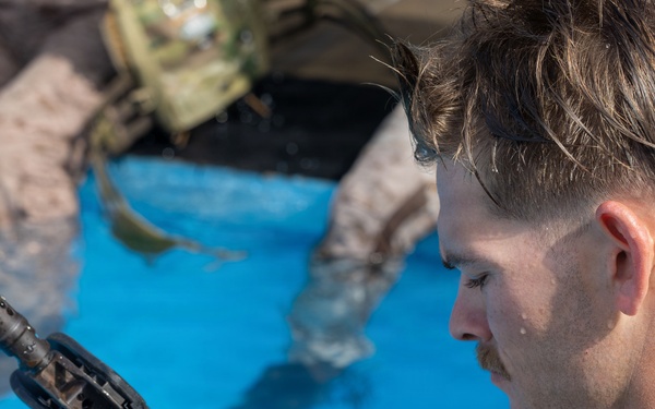 A military service member in desert MARPAT camouflage uniform is inspecting or field-stripping a firearm component while partially submerged in water, likely during a water survival or amphibious training exercise. A second service member in similar uniform is visible in the blurred background, also in the water. The setting appears to be a training pool based on the bright blue water visible.
