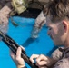 A military service member in desert MARPAT camouflage uniform is inspecting or field-stripping a firearm component while partially submerged in water, likely during a water survival or amphibious training exercise. A second service member in similar uniform is visible in the blurred background, also in the water. The setting appears to be a training pool based on the bright blue water visible.