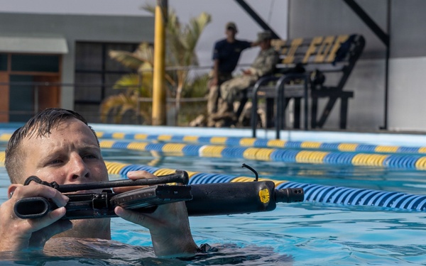 A person is submerged chest-deep in a swimming pool while holding what appears to be a military-style rifle or weapon above the waterline. The setting appears to be a formal training exercise, as observers in military-style clothing are visible seated in the background. Lane dividers in blue and yellow are visible across the pool.