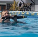 A person is submerged chest-deep in a swimming pool while holding what appears to be a military-style rifle or weapon above the waterline. The setting appears to be a formal training exercise, as observers in military-style clothing are visible seated in the background. Lane dividers in blue and yellow are visible across the pool.