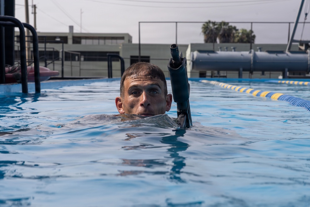 A male swimmer is partially submerged in an outdoor lap pool, with only his head and shoulders visible above the water surface. He is wearing a wetsuit and has a snorkel attached near his head, suggesting diving or underwater training activity. The setting appears to be a military or institutional facility based on the infrastructure visible in the background.