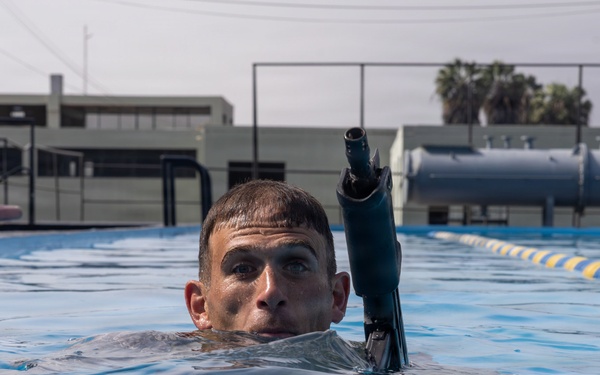 A male swimmer is partially submerged in an outdoor lap pool, with only his head and shoulders visible above the water surface. He is wearing a wetsuit and has a snorkel attached near his head, suggesting diving or underwater training activity. The setting appears to be a military or institutional facility based on the infrastructure visible in the background.