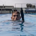 A male swimmer is partially submerged in an outdoor lap pool, with only his head and shoulders visible above the water surface. He is wearing a wetsuit and has a snorkel attached near his head, suggesting diving or underwater training activity. The setting appears to be a military or institutional facility based on the infrastructure visible in the background.