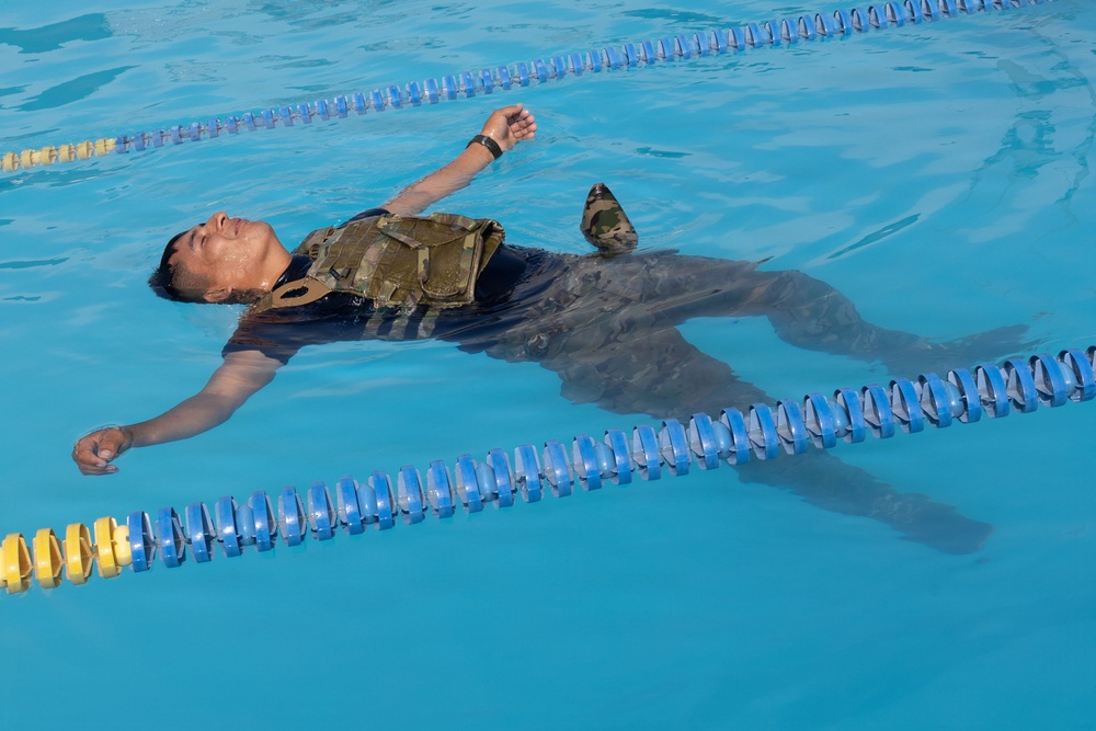 A person is floating on their back in a swimming pool while wearing full military/tactical gear including a camouflage plate carrier vest and combat boots. The individual appears to be conducting a military or law enforcement water survival training exercise. The pool is a standard lap pool with blue and yellow lane dividers.