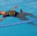A person is floating on their back in a swimming pool while wearing full military/tactical gear including a camouflage plate carrier vest and combat boots. The individual appears to be conducting a military or law enforcement water survival training exercise. The pool is a standard lap pool with blue and yellow lane dividers.