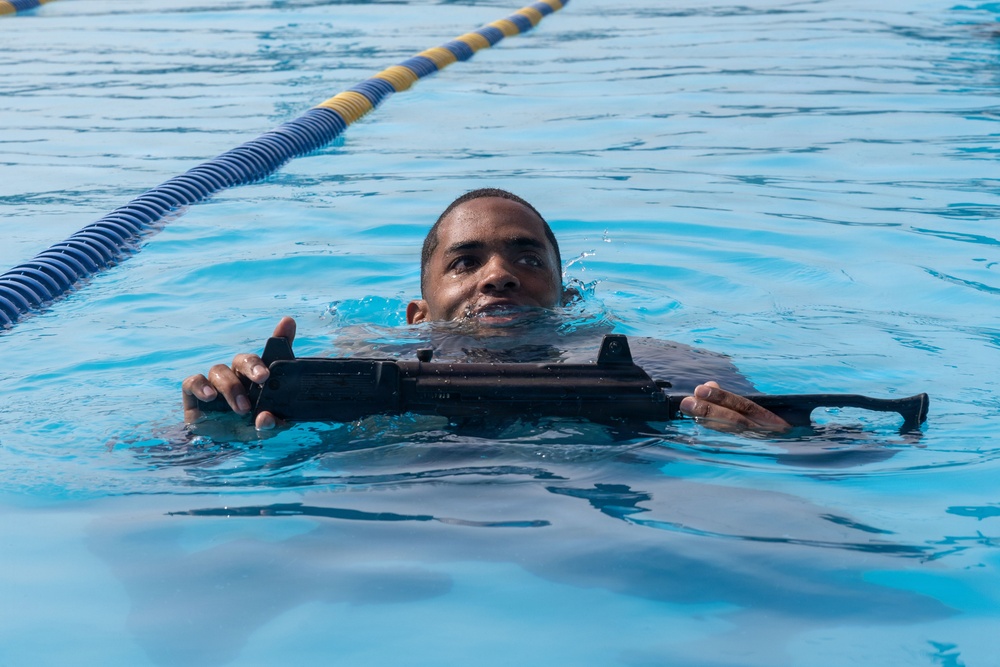 A person is swimming in a marked lap pool while holding a rifle across their chest. The individual appears to be participating in military or law enforcement aquatic training, keeping their head above water while maintaining control of the weapon. The setting is a structured swimming facility with lane dividers visible in the background.