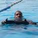 A person is swimming in a marked lap pool while holding a rifle across their chest. The individual appears to be participating in military or law enforcement aquatic training, keeping their head above water while maintaining control of the weapon. The setting is a structured swimming facility with lane dividers visible in the background.