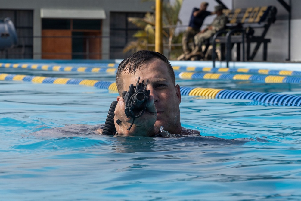 A military or law enforcement trainee is partially submerged in a swimming pool while aiming a rifle with an optic sight directly at the camera. The setting appears to be a military or tactical training facility with lane markers visible in the pool. Additional personnel in military/tactical attire are visible seated in the background.