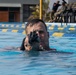 A military or law enforcement trainee is partially submerged in a swimming pool while aiming a rifle with an optic sight directly at the camera. The setting appears to be a military or tactical training facility with lane markers visible in the pool. Additional personnel in military/tactical attire are visible seated in the background.
