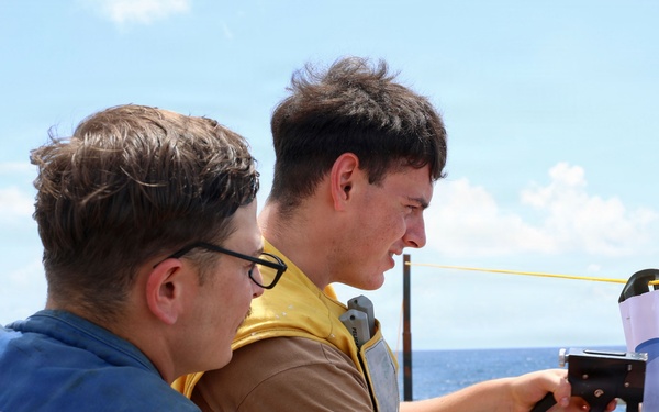 Two military personnel are working together on the deck of a naval vessel, identified by the 'DDG 113' marking on a blue vest. One sailor is using a staple gun or similar tool to attach a paper or document to a surface, while the other observes closely. The scene takes place at sea under bright, partly cloudy skies.
