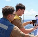 Two military personnel are working together on the deck of a naval vessel, identified by the 'DDG 113' marking on a blue vest. One sailor is using a staple gun or similar tool to attach a paper or document to a surface, while the other observes closely. The scene takes place at sea under bright, partly cloudy skies.