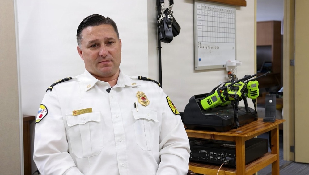 A uniformed fire department official, likely a chief or senior officer, is seated in what appears to be a fire station administrative office or communications room. He is wearing a white dress uniform with gold badge, rank insignia, and department patches. Behind him are radio charging stations with neon green portable radios, a whiteboard, and communications equipment.