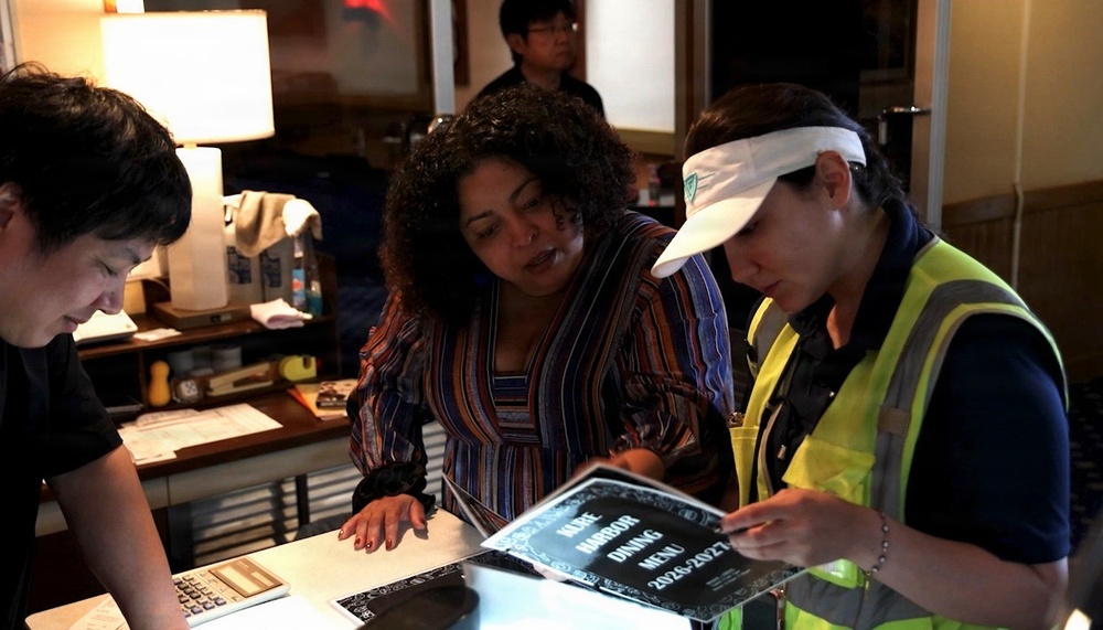 Three people are gathered around a counter or desk in what appears to be a dimly lit restaurant or dining facility. A woman in a yellow safety vest and white visor is holding a dark menu booklet labeled 'Harbor Dining Menu 2026-2027', while a woman in a striped blouse leans in to look at it. A fourth person is visible in the background.