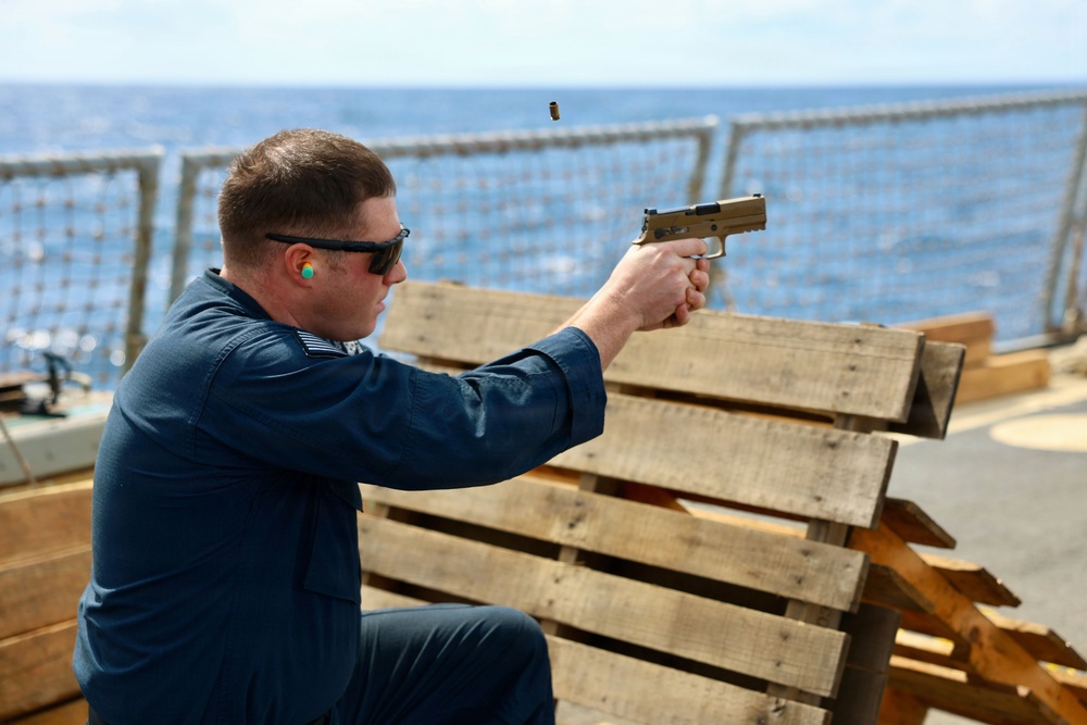 A person in a blue uniform is firing a tan/flat dark earth semi-automatic pistol from a shooting position on what appears to be the deck of a naval vessel. A spent brass casing is visibly ejecting from the firearm. The shooter is using wooden pallets as a shooting rest and is wearing ear protection and safety glasses.