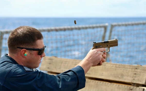A person in a blue uniform is firing a tan/flat dark earth semi-automatic pistol from a shooting position on what appears to be the deck of a naval vessel. A spent brass casing is visibly ejecting from the firearm. The shooter is using wooden pallets as a shooting rest and is wearing ear protection and safety glasses.