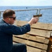 A person in a blue uniform is firing a tan/flat dark earth semi-automatic pistol from a shooting position on what appears to be the deck of a naval vessel. A spent brass casing is visibly ejecting from the firearm. The shooter is using wooden pallets as a shooting rest and is wearing ear protection and safety glasses.