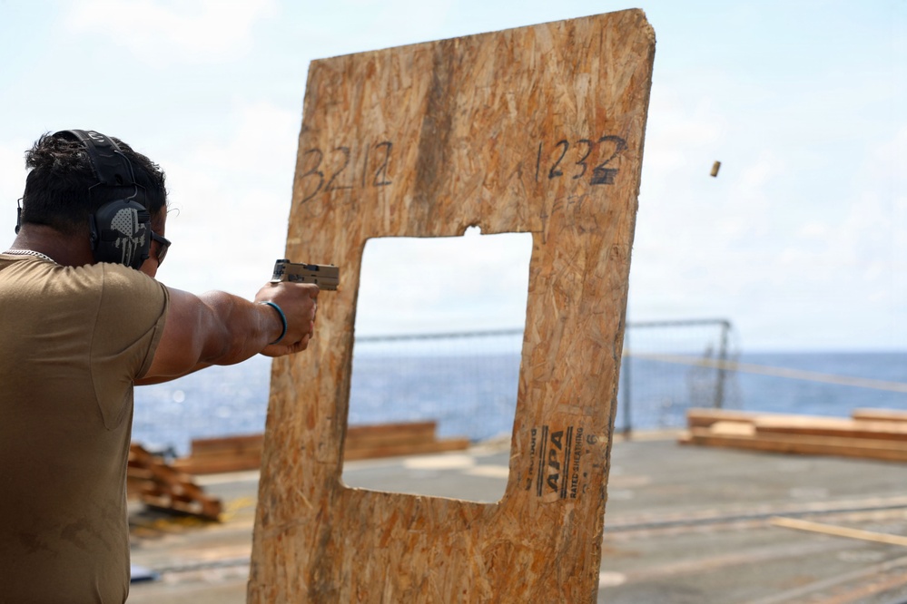 A person wearing hearing protection and safety glasses is firing a pistol at a plywood target board on what appears to be the deck of a naval vessel at sea. A spent brass casing is visibly ejecting from the firearm. The setting suggests a shipboard firearms qualification or training exercise.