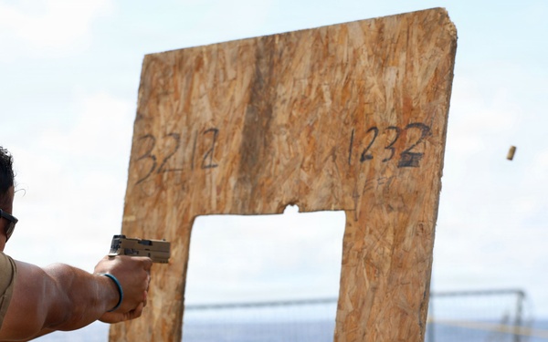 A person wearing hearing protection and safety glasses is firing a pistol at a plywood target board on what appears to be the deck of a naval vessel at sea. A spent brass casing is visibly ejecting from the firearm. The setting suggests a shipboard firearms qualification or training exercise.