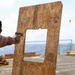 A person wearing hearing protection and safety glasses is firing a pistol at a plywood target board on what appears to be the deck of a naval vessel at sea. A spent brass casing is visibly ejecting from the firearm. The setting suggests a shipboard firearms qualification or training exercise.