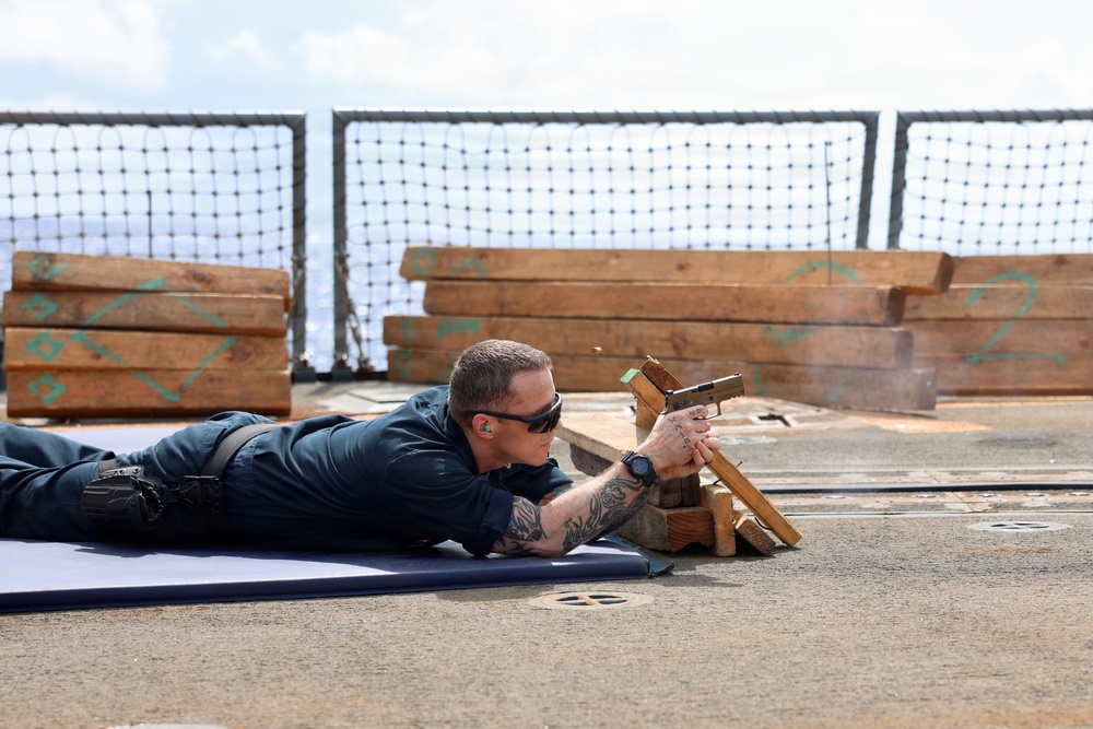 A person in dark navy/blue uniform is lying prone on a blue mat on what appears to be a ship's deck, aiming a pistol over a wooden rest. The setting includes stacked lumber with green markings and a safety net barrier in the background, consistent with an improvised shipboard firing range. The shooter is wearing ear protection, sunglasses, and has visible tattoos on both forearms.