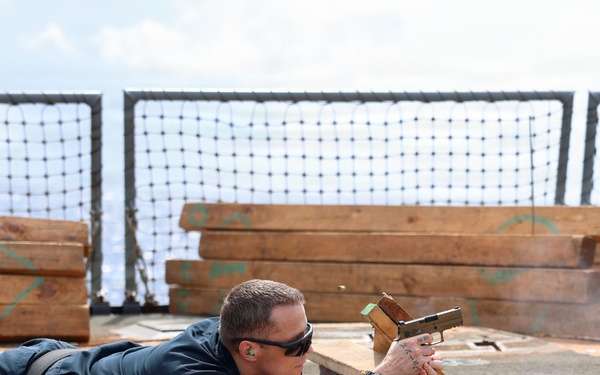 A person in dark navy/blue uniform is lying prone on a blue mat on what appears to be a ship's deck, aiming a pistol over a wooden rest. The setting includes stacked lumber with green markings and a safety net barrier in the background, consistent with an improvised shipboard firing range. The shooter is wearing ear protection, sunglasses, and has visible tattoos on both forearms.